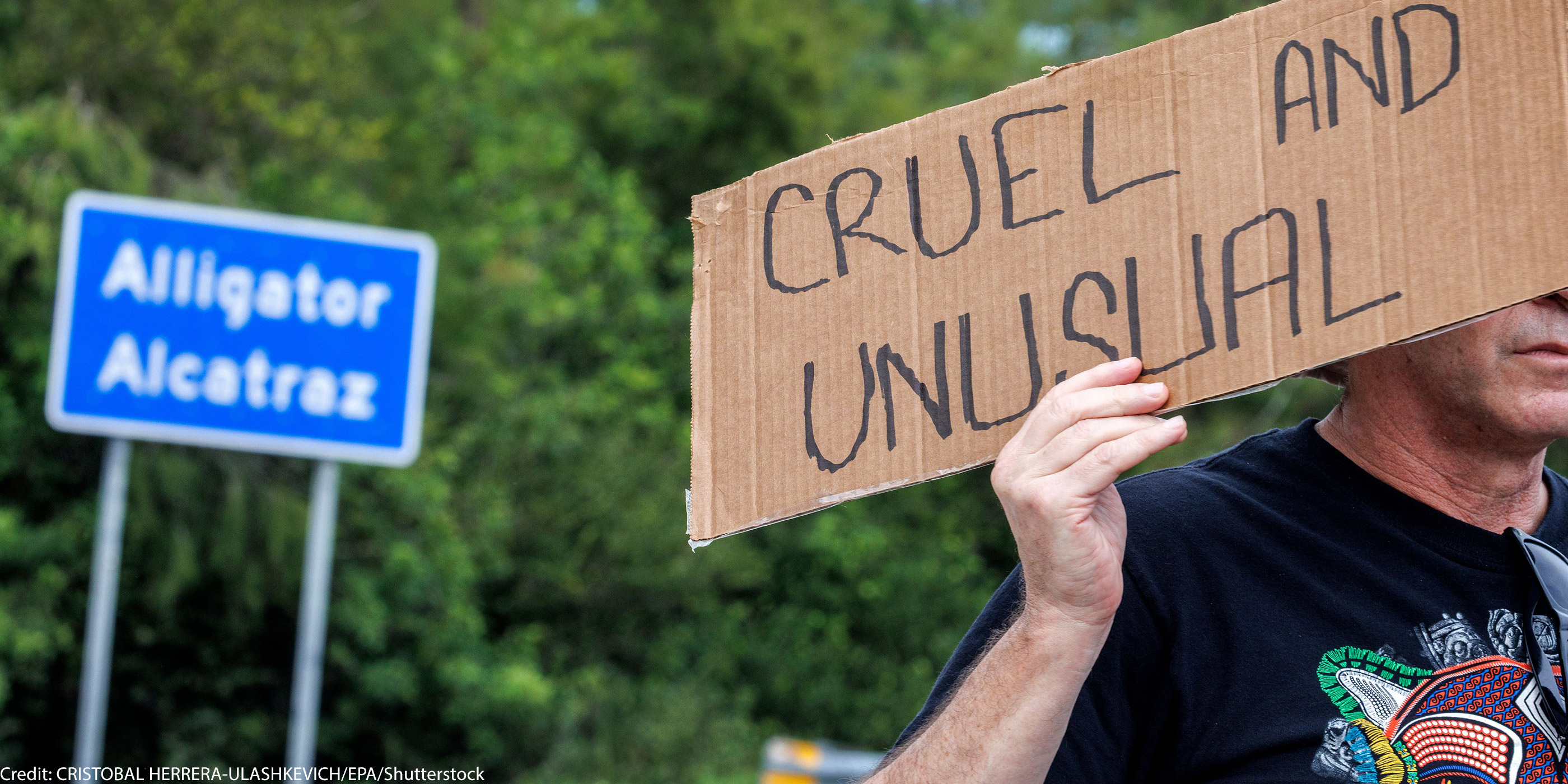 A road sign that says Alligator Alcatraz and an individual in front of it holding a sign that says "Cruel and Unusual."