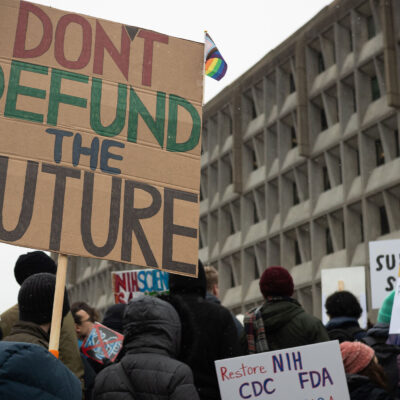 People attend a rally against the Trump administration's 90-day funding freeze and job cuts at health agencies in Washington, DC, hold up a large sign written in red, green and black on cardboard that reads " DON'T DEFUND THE FUTURE."