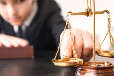 A child dressed in a suit with a hand on top of a book, tips scales of justice.