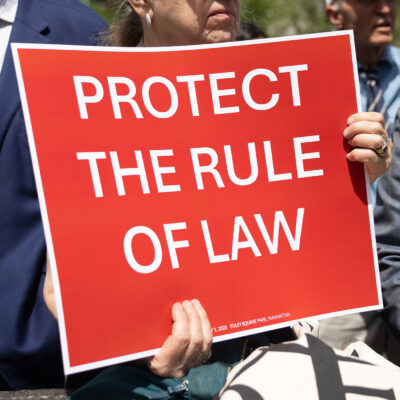 At the Lawyers Rally In Lower Manhattan To Demand Respect For The Rule Of Law, a demonstrator holds up a sign with white lettering on a red background that reds, "PROTECT THE RULE OF LAW".