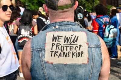 At a trans rally, person (wearing a denim vest and with their back to the camera) displays the sign stitched to the vest that reads "WE WILL PROTECT TRANS KIDS."