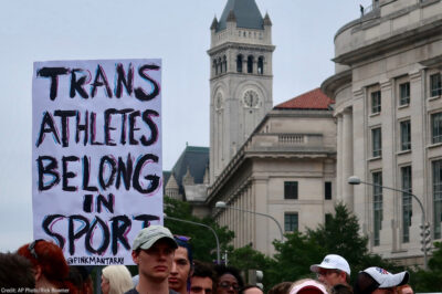 A crowd of marchers with one holding a sign with the text " Trans Athletes Belong in Sport."